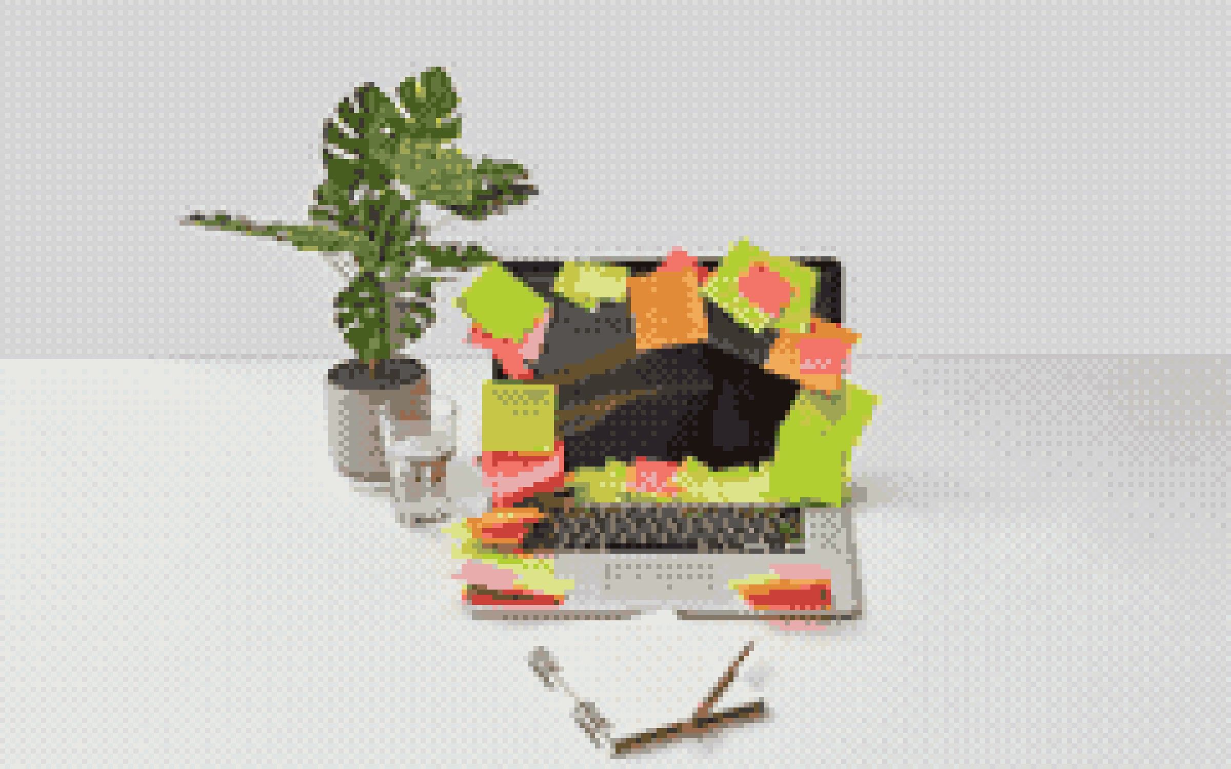 A laptop covered in colorful sticky-note reminders beside a plant, glass of water, and open notebook.